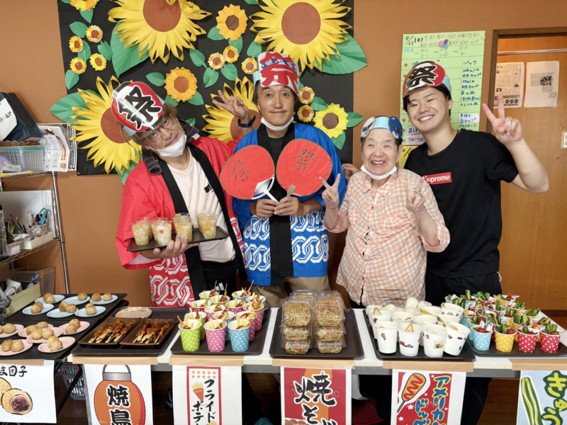 夏祭りの屋台越しに記念撮影をする利用者さまたちとスタッフたち