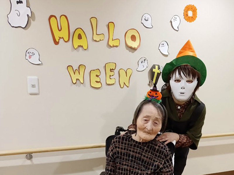 ハロウィンの壁飾りの前でスタッフと一緒に写真に写った入居者さま