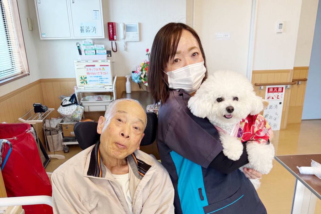 入居者さまのそばでメルちゃんを抱っこするスタッフ