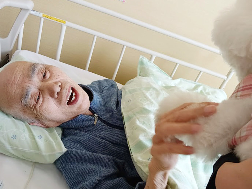 ベッドからメルちゃんの足に触れて笑顔の入居者さま